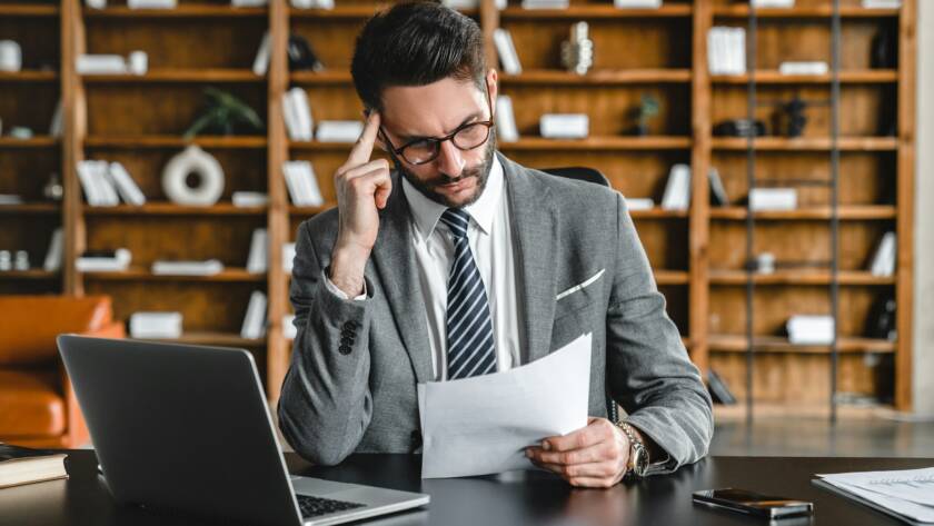 Concentrated young foreman reading legal papers while working in office on laptop